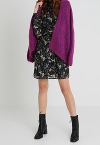 Schwarzes Blumenmuster-Kleid mit langen Ärmeln und taillierter Taille, kombiniert mit einer groben lila Strickjacke. Schwarze Schnürstiefeletten runden den Look ab.
