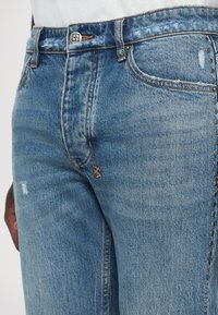 Gros plan sur un jean en denim bleu avec des clous métalliques le long de la couture latérale extérieure et un léger effet usé près de la poche et de la cuisse.