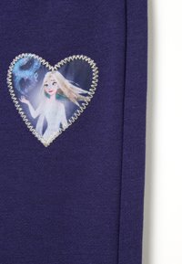 Paarse katoenen top met een hartvormige patch met een geprinte karakterafbeelding, omrand met zilveren steken, op een gestructureerde stof.