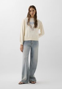 Creme sweatshirt med grafisk tryk, der forestiller en ørn, parret med lyseblå flared jeans. Flip-flops fuldender det afslappede look.