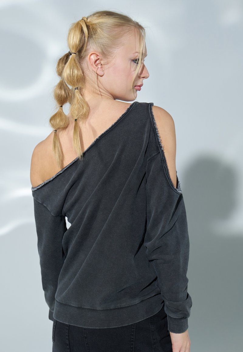 Felpa nera off-shoulder con bordi grezzi, maniche lunghe e orlo a costine. Indossata sopra pantaloni scuri, mostra un design casual e rilassato.