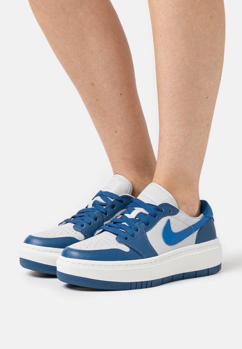Paire de baskets Nike blanches et bleues avec des semelles épaisses portées pieds nus sur fond blanc.