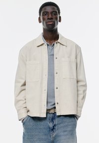 Lysbeige jakke med to forlommer, knaplukning og krave, lagd over en grå polo-shirt, kombineret med blå denim.
