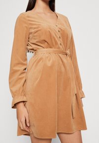 Donna che indossa un abito di velluto a coste lungo, color beige, con maniche lunghe, bottoni frontali e una cintura legata in vita, su uno sfondo semplice.