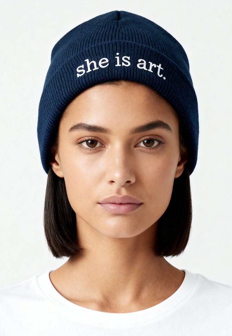 Bonnet en tricot bleu marine avec une texture côtelée, arborant le texte « she is art. » brodé en blanc sur le devant. Porté avec un T-shirt blanc.