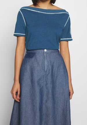 Kvinde iført en blågrøn off-shoulder kortærmet top med hvide syninger og en højtaljet blå A-line nederdel med knap- og lomme detaljer.