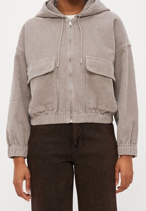 Lichtgrijze corduroy cropped hoodie met voorsluiting met rits, twee grote klepzakken en elastische manchetten, gedragen met donkerbruine broek.