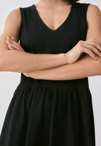 Sort ærmeløs kjole med V-udskæring, indsnævret talje og udvidet nederdel. Stoffet virker blødt med en glat tekstur. Enkel design.