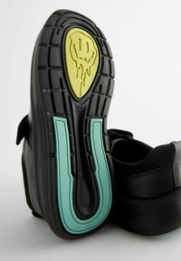 Chaussure noire avec une semelle en caoutchouc texturée présentant un design de visage souriant jaune et des accents turquoise le long de la semelle. Boucle Velcro visible.