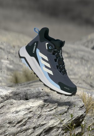 Dark gray hiking shoe with white and light blue accents, textured upper, sturdy sole, and raised "Terrex" logo, suspended above rocky terrain.