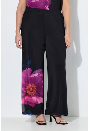 Vrouw draagt zwarte wijde broek met grote roze en oranje bloemenprint op één pijp en zwarte sandalen met hak.