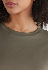 Olivgrüner, strukturierter Stoff mit Rundhalsausschnitt, der über glatte Nähte und ein enganliegendes Design verfügt. Haut ist am Ausschnitt sichtbar.
