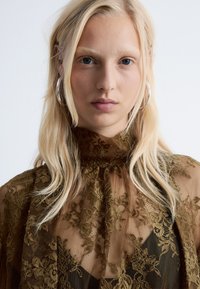 Durchsichtige olivgrüne Spitzentop mit hohem Kragen, floralen Mustern und transparenter Textur. Silberne Creolen setzen einen dezenten metallischen Akzent.