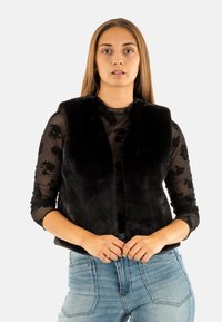 Chaleco sin mangas de pelaje sintético negro con cuello de chal, usado sobre una blusa negra transparente de manga larga con patrones florales, combinado con jeans azules.