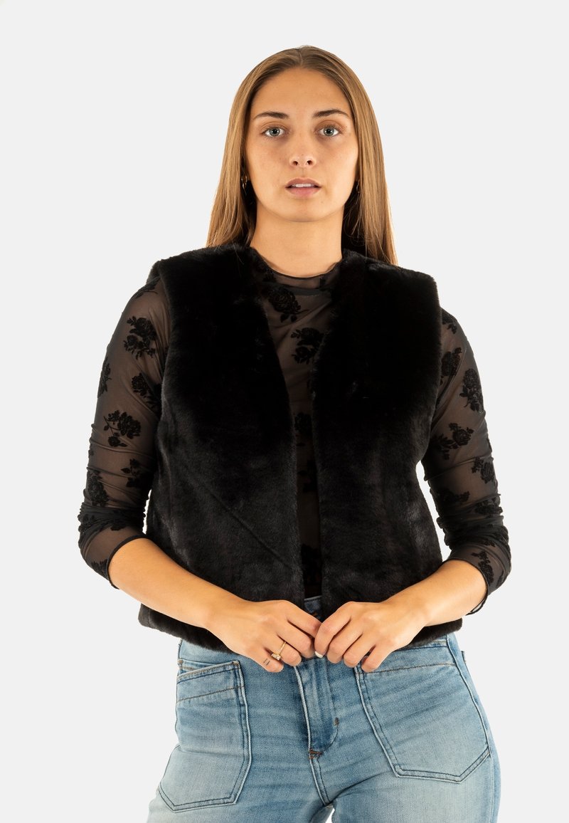 Chaleco sin mangas de pelaje sintético negro con cuello de chal, usado sobre una blusa negra transparente de manga larga con patrones florales, combinado con jeans azules.