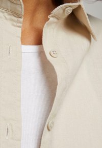 Gros plan d’une personne portant un t-shirt blanc côtelé sous une chemise beige boutonnée avec boutons visibles et détails du col.