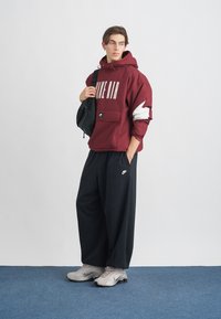 Sweatshirt com capuz maroon e logótipo branco, calças largas pretas e sapatilhas Nike cinzas claras. O acessório inclui uma mochila preta.