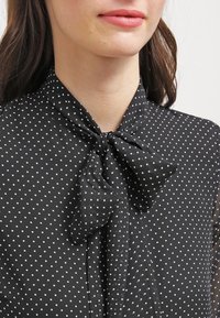 Blusa nera con pois bianchi, caratterizzata da un dettaglio con fiocco al collo. Il tessuto ha una texture liscia con un aspetto semi-trasparente.