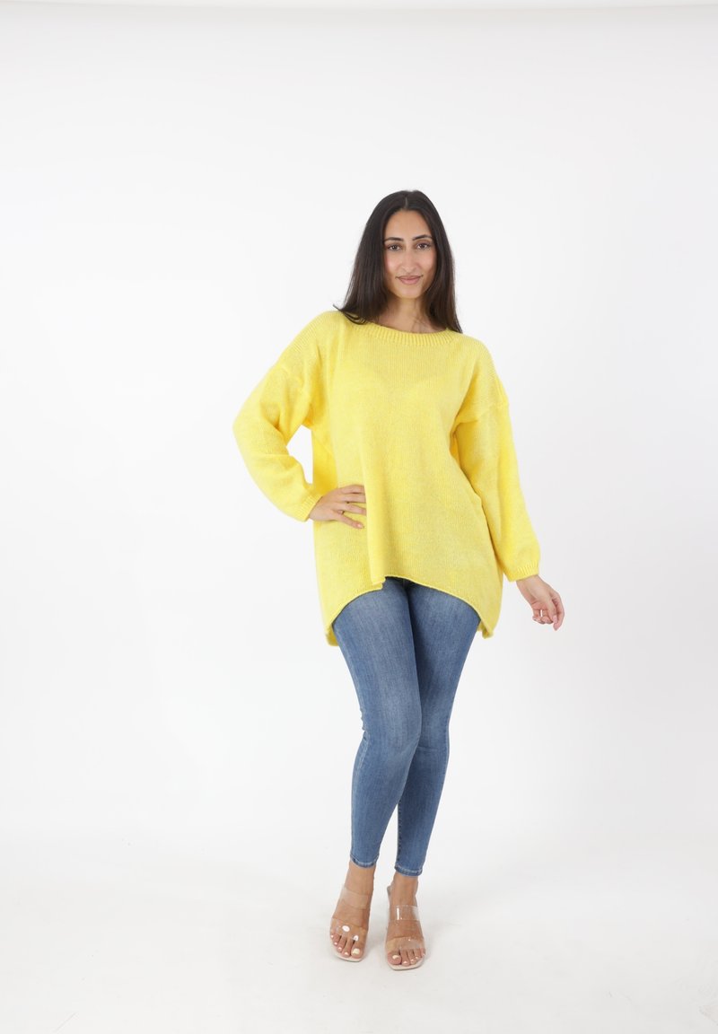 Żółty, dzianinowy, oversize'owy sweter z okrągłym dekoltem, długimi rękawami i asymetrycznym dołem; w połączeniu z niebieskimi, wąskimi dżinsami i przezroczystymi sandałami na obcasie.