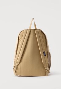 Μπεζ σακίδιο πλάτης Jansport, εμφανιζόμενο από πίσω, με επενδεδυμένους ιμάντες ώμου, δίχτυα πλαϊνές θήκες και θηλιά μεταφοράς στην κορυφή.