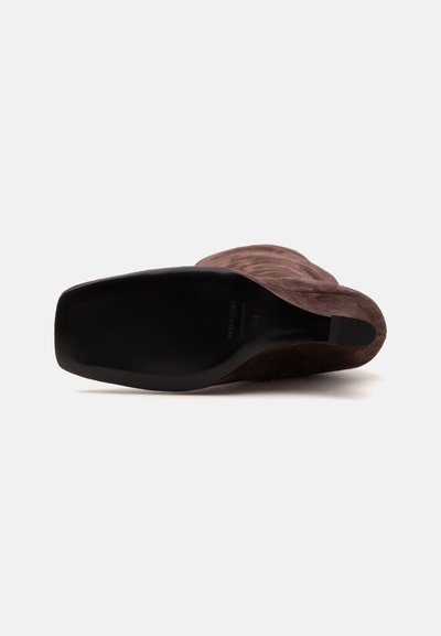 Botte haute au genou en daim marron, couchée sur le côté, montrant une semelle noire à bout carré avec un petit talon sur fond blanc.