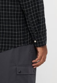 Camisa xadrez preta e branca com mangas compridas e punhos com botões, combinada com calças cinzentas com bolsos laterais e fechos de encaixe.