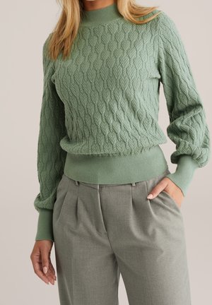 Femme portant un pull en maille vert sauge texturé avec des manches bouffantes et un pantalon plissé taille haute vert olive, une main dans la poche.