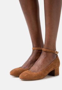 Décolleté in suede marrone con punta arrotondata e tacco block di media altezza, dotate di cinturino alla caviglia con una piccola fibbia dorata.