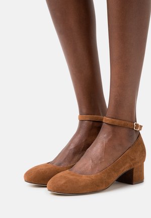 Décolleté in suede marrone con punta arrotondata e tacco block di media altezza, dotate di cinturino alla caviglia con una piccola fibbia dorata.