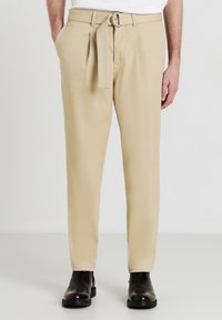 Pantaloni beige realizzati in tessuto testurizzato con dettaglio della cintura a anello e una vestibilità dritta. Presentano tasche laterali e una piega sul davanti.