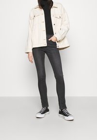 Chaqueta de denim crema sobre un cuello de tortuga negro, combinada con jeans ajustados gris oscuro y zapatillas negras de caña alta con suelas blancas.