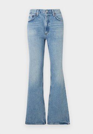 Femme debout, les mains dans les poches, portant un haut noir à manches courtes avec un liseré blanc, un jean bleu clair droit et des chaussures beige pointues.