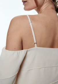 Leichtes, schulterfreies Oberteil in Creme mit dünnen Trägern und einer gesteppten Verzierung. Die glatte Textur und die lockere Passform betonen den Rücken.