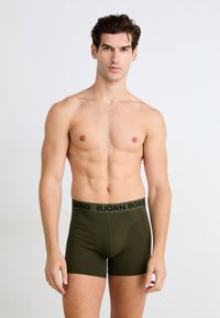 Olivengrønne boxershorts med en sort elastisk talje, der har "BJÖRN BORG"-logoet. Glat tekstur og tætsiddende design.