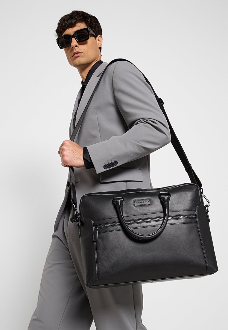 Malette en cuir noir avec deux poignées supérieures et une bandoulière amovible. Présente un poche zippée à l'avant et une texture lisse, portée par un mannequin.