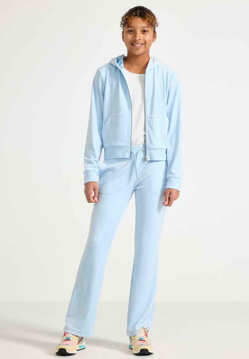 Jasnoniebieska bluza z kapturem na zamek z przednimi kieszeniami, zestawiona z dopasowanymi dresami. Miękka tekstura materiału i elastyczny pas ze sznurkiem do ściągania. Sneakersy w kolorze dopasowanym do stroju.
