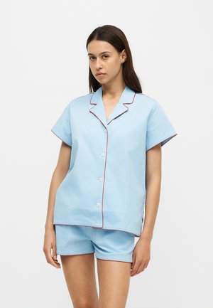 Vrouw draagt een lichtblauw kort mouw knoophemd en bijpassende korte broek met rode bies, staand tegen een egale witte achtergrond.