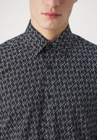 Camicia nera con bottoni caratterizzata da un motivo ripetitivo di testo bianco scritto a mano, con colletto strutturato e texture di tessuto morbido.