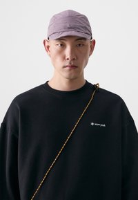 Casquette violette avec des panneaux cousus et un logo sur le devant, associée à un sweat-shirt noir arborant un logo blanc et une sangle tressée.