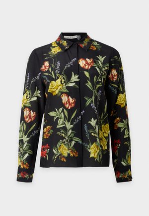 Femme portant une chemise noire à fleurs à manches longues, un jean bleu à jambes larges et des chaussures noires pointues, debout devant un fond uni.