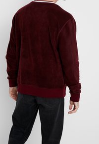 Bordeaux fleece sweatshirt med ribbede manchetter og kant, med en rund halsudskæring og en glat tekstur. Bæres med mørke jeans.