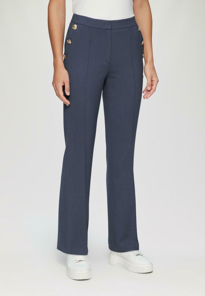 Pantalons navy ajustés avec quatre boutons dorés sur le côté, design à jambes droites et texture lisse. Associés à des baskets blanches.