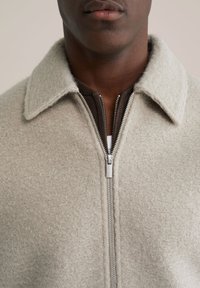 Jachetă din lână gri deschis cu finisaj texturat, având guler și fermoar argintiu. Purta peste un articol vestimentar maro închis dedesubt.