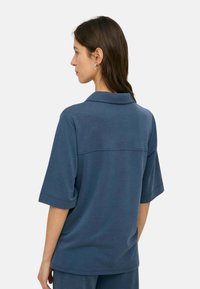 Mujer con cabello largo y castaño, vestida con una camiseta polo azul de manga corta y suelta y pantalones a juego, de perfil contra un fondo blanco.