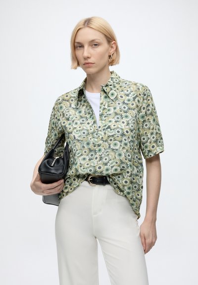 Woman with short blonde hair wearing a green floral short-sleeve shirt, white pants, holding a black handbag against a plain background.