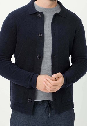 Mann trägt marineblauen Cardigan mit Knöpfen über hellgrauem geripptem Pullover und dunkler, strukturierter Hose, die Hände vor der Taille verschränkt.