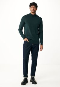 Tmavě zelený rolák vyrobený z jemného pleteného materiálu, doplněný o tmavě modré džíny a černé kotníkové boty s masivní podrážkou.