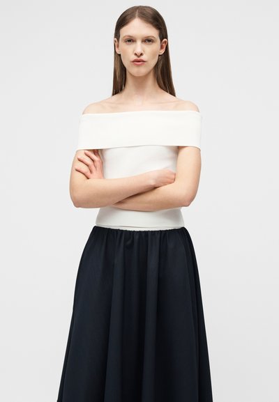 Femme aux cheveux longs bruns, vêtue d'un haut blanc épaules dénudées et d'une jupe noire, debout les bras croisés devant un fond clair uni.