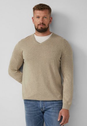 Beżowy sweter w serek z miękkiego dzianiny, z długimi rękawami i dopasowanymi mankietami, w zestawieniu z niebieskimi dżinsami. Minimalistyczny detal z logo na klatce piersiowej.