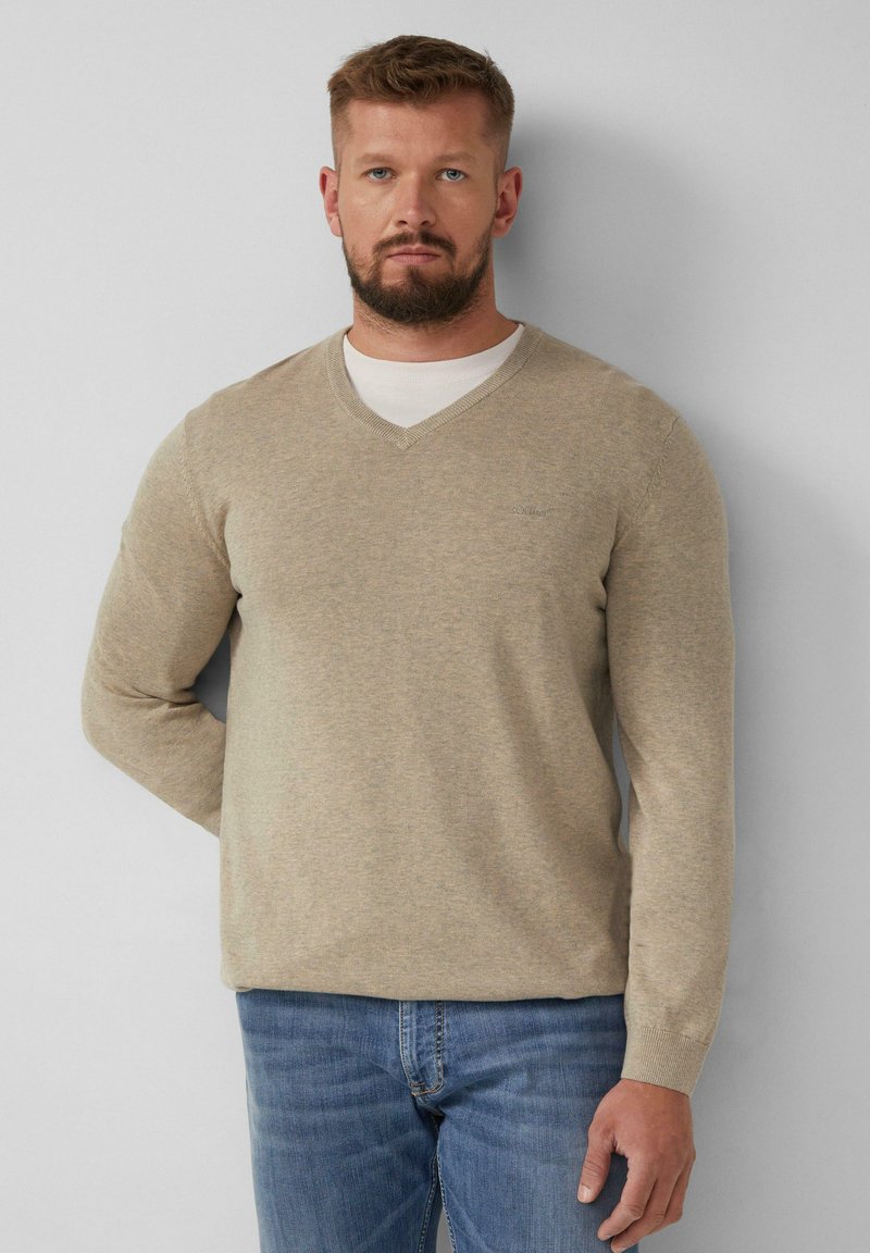 Beige V-Ausschnitt-Pullover aus einem weichen Strickmaterial, mit langen Ärmeln und eng anliegenden Bündchen, kombiniert mit blauen Jeans. Minimales Branding-Detail auf der Brust.